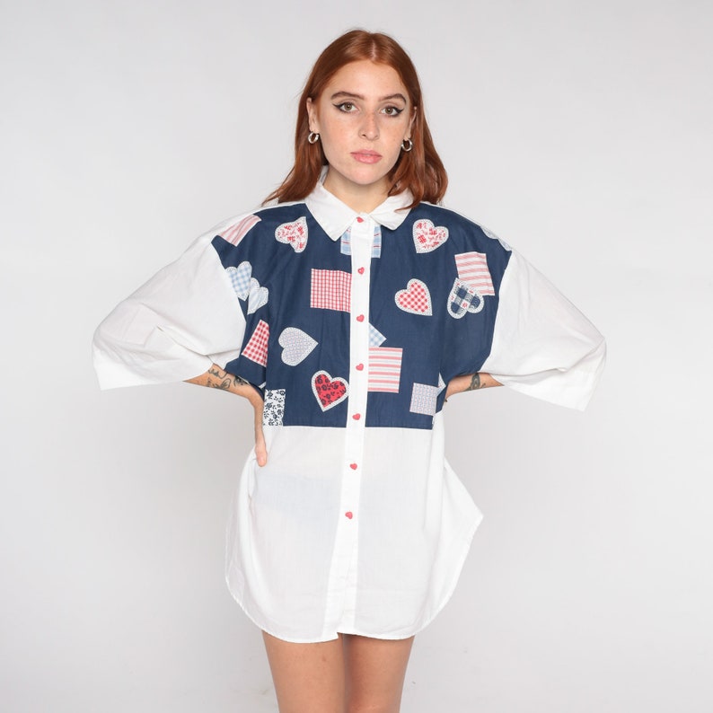 May include: A white button-down shirt with a blue panel featuring a patchwork design of red and white hearts and squares.