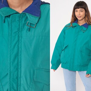 K&ouml;nnte beinhalten: Eine tealblaue Kapuzenjacke mit Rei&szlig;verschluss und zwei Fronttaschen. Die Jacke hat ein marineblaues Futter um die Kapuze.
