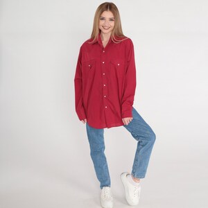 May include: A red long-sleeved button-down shirt with a collared neckline and two chest pockets. The shirt is worn with blue jeans and white trainers.