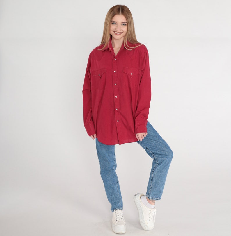May include: A red long-sleeved button-down shirt with a collared neckline and two chest pockets. The shirt is worn with blue jeans and white trainers.