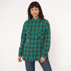 May include: A green and black plaid flannel shirt with two chest pockets and a button-down front. The long-sleeved shirt is made of a soft, textured fabric. The shirt is paired with blue jeans.