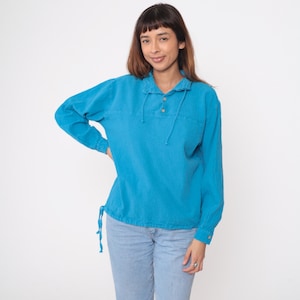 Pode incluir: Uma camisa turquesa de manga comprida com gola e cordão. A camisa tem dois botões e um cordão no decote e na cintura. A modelo está vestida com calças de ganga azul clara.