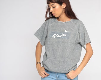 May include: Gray short-sleeved sweatshirt with a white graphic of a bird flying over a grid pattern and the word "Alaska" written in cursive.