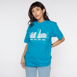 May include: A turquoise t-shirt featuring a white graphic of the Seattle skyline, including the Space Needle. The text "DATA COMPUTER ARCHITECTURE SEATTLE 1990" is printed below the skyline design. The shirt has short sleeves.