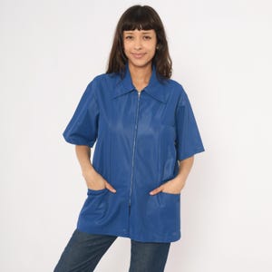 May include: A short-sleeved, cobalt blue zip-up jacket with a collar and two front pockets. The jacket is made of a smooth, possibly synthetic material. The model is wearing blue jeans.