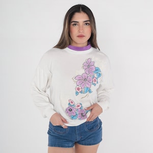 May include: White long-sleeved shirt with a floral print featuring pink, purple and blue flowers. The shirt has a crew neck and a relaxed fit.