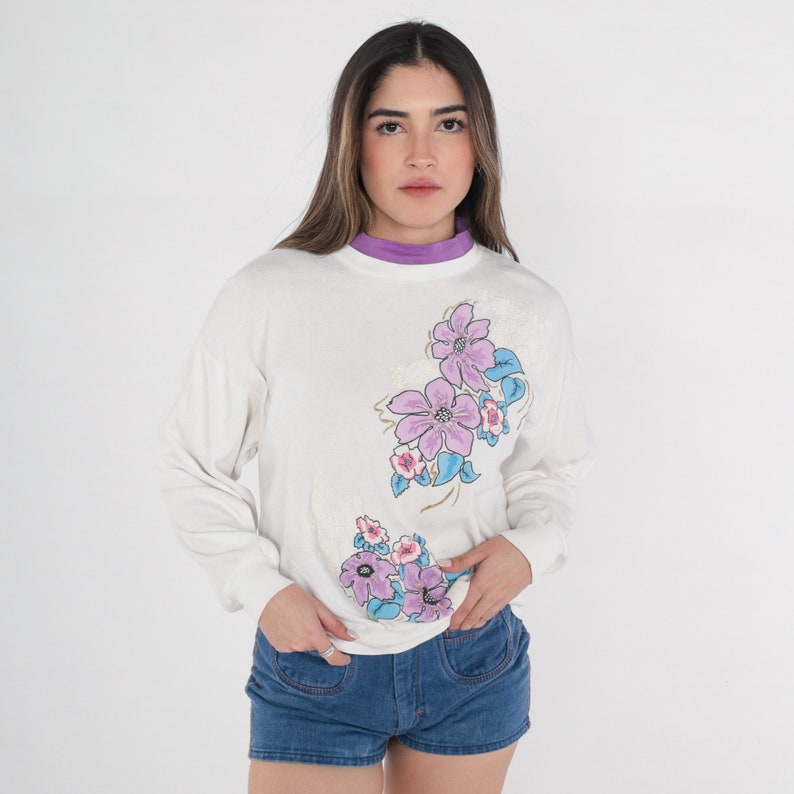 May include: White long-sleeved shirt with a floral print featuring pink, purple and blue flowers. The shirt has a crew neck and a relaxed fit.