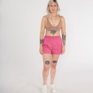 May include: A woman wearing a tan-coloured bra and hot pink shorts. The shorts have an elastic waistband. She is also wearing white platform trainers and a necklace. The background is plain white.