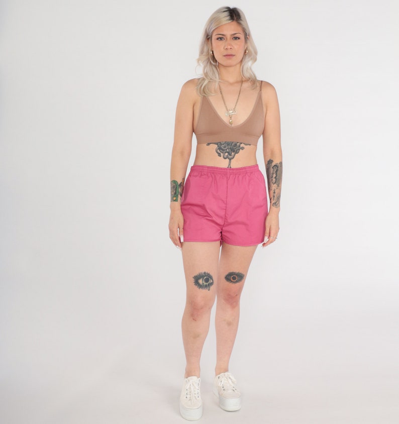 May include: A woman wearing a tan-coloured bra and hot pink shorts. The shorts have an elastic waistband. She is also wearing white platform trainers and a necklace. The background is plain white.