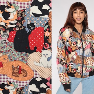 Puede incluir: Chaqueta bomber colorida con un diseño de patchwork con varias ilustraciones de gatos. La chaqueta tiene cuello, puños y dobladillo negros, y cierre de cremallera. Los patrones de gatos incluyen gatos calicó, negros y naranjas, con detalles florales y de lunares.