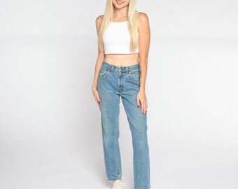 May include: A woman wearing a white crop top and light blue denim jeans. She is standing with her feet slightly apart and her hands at her sides. She is smiling at the camera.