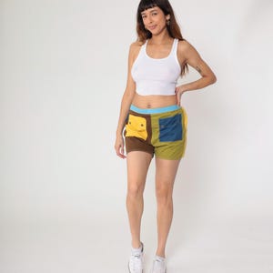 May include: A woman wearing a white tank top and colorful patchwork shorts. The shorts feature brown, yellow, blue, and green panels with pockets. The shorts have a blue waistband.