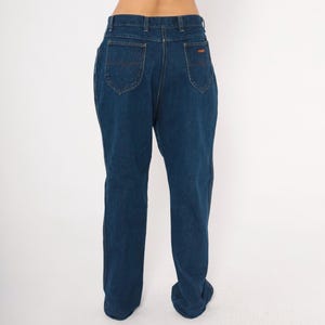 May include: Dark blue denim jeans with a classic five-pocket design. The jeans have a straight leg cut and a high waist. The back pockets feature a curved stitch detail. A small brown label is visible on the back waistband.
