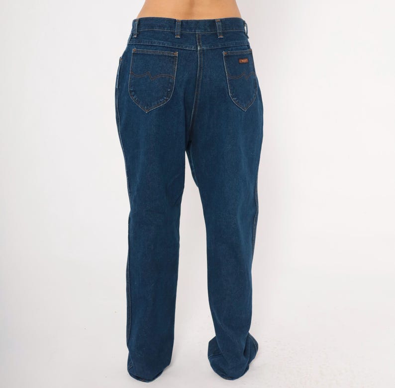 May include: Dark blue denim jeans with a classic five-pocket design. The jeans have a straight leg cut and a high waist. The back pockets feature a curved stitch detail. A small brown label is visible on the back waistband.