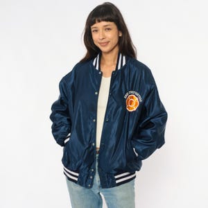 May include: Navy blue satin bomber jacket with white and black striped trim on the collar, cuffs, and hem. The jacket features a Firefighters emblem on the chest. The jacket has a button-up front.