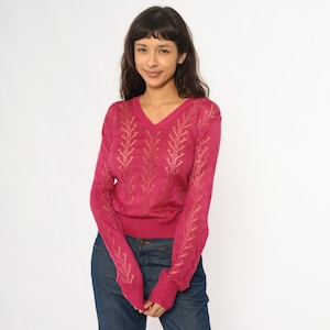 May include: A magenta-colored, long-sleeved sweater with a V-neck. The sweater features a leaf-like openwork design down the front and sleeves. The person in the image is wearing blue jeans.