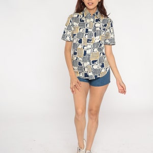 May include: A white short-sleeved button-down shirt with a geometric pattern in blue and yellow. The shirt is worn with denim shorts and white sneakers.