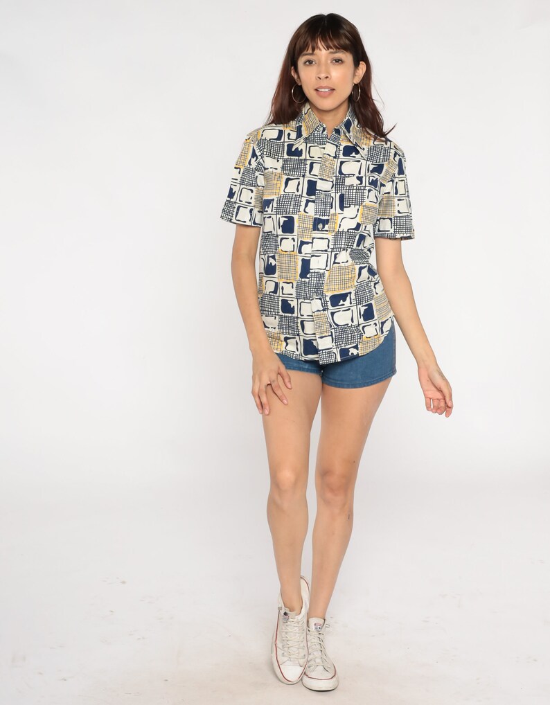 May include: A white short-sleeved button-down shirt with a geometric pattern in blue and yellow. The shirt is worn with denim shorts and white sneakers.