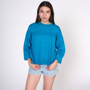 May include: A teal blue sweatshirt with a horizontal stripe across the chest. The sweatshirt has a relaxed fit and a crew neck.