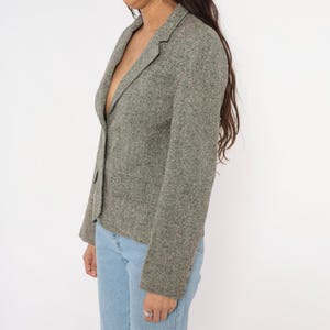 May include: A gray tweed blazer with a single-button closure and two front pockets. The blazer is worn with light blue jeans. The jacket has a classic, tailored design.