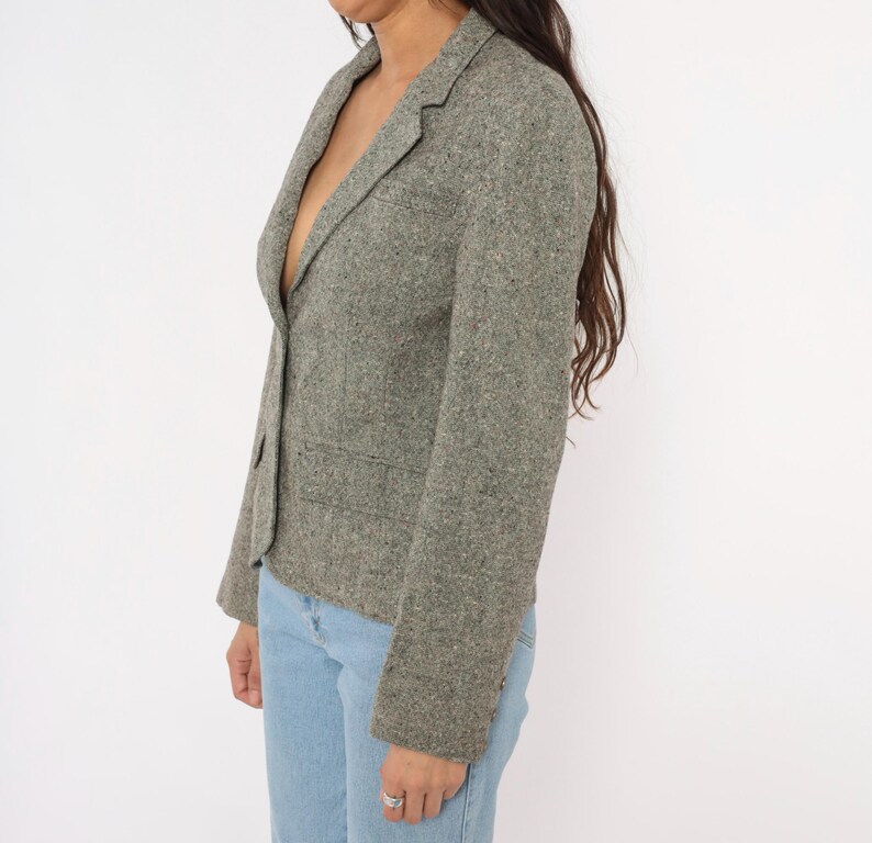 May include: A gray tweed blazer with a single-button closure and two front pockets. The blazer is worn with light blue jeans. The jacket has a classic, tailored design.
