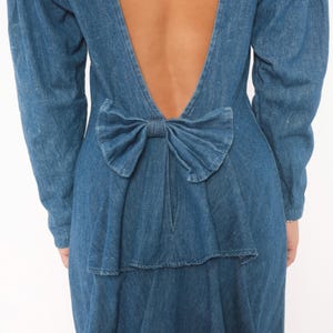 Puede incluir: Un vestido de mezclilla azul de manga larga con un dise&ntilde;o de espalda abierta en forma de V profunda. La prenda est&aacute; acentuada con un lazo grande de mezclilla fruncido colocado en la parte baja de la espalda, justo encima de la cintura.