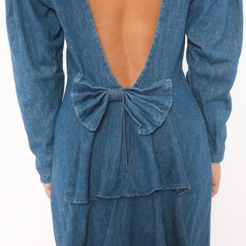 Puede incluir: Un vestido de mezclilla azul de manga larga con un dise&ntilde;o de espalda abierta en forma de V profunda. La prenda est&aacute; acentuada con un lazo grande de mezclilla fruncido colocado en la parte baja de la espalda, justo encima de la cintura.