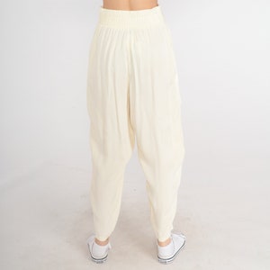 May include: Cream-colored, loose-fitting pants with an elastic waistband. The pants have a relaxed, comfortable style, and are paired with white sneakers. The fabric appears to be lightweight and flowing, suitable for casual wear.