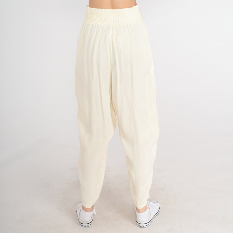May include: Cream-colored, loose-fitting pants with an elastic waistband. The pants have a relaxed, comfortable style, and are paired with white sneakers. The fabric appears to be lightweight and flowing, suitable for casual wear.