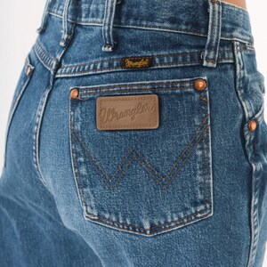 Puede incluir: Primer plano de unos vaqueros azules con una etiqueta Wrangler en el bolsillo trasero. Los vaqueros tienen un dise&ntilde;o cl&aacute;sico de cinco bolsillos con remaches de cobre y un parche de cuero marr&oacute;n. El logotipo de Wrangler es visible en el parche y encima del bolsillo trasero.