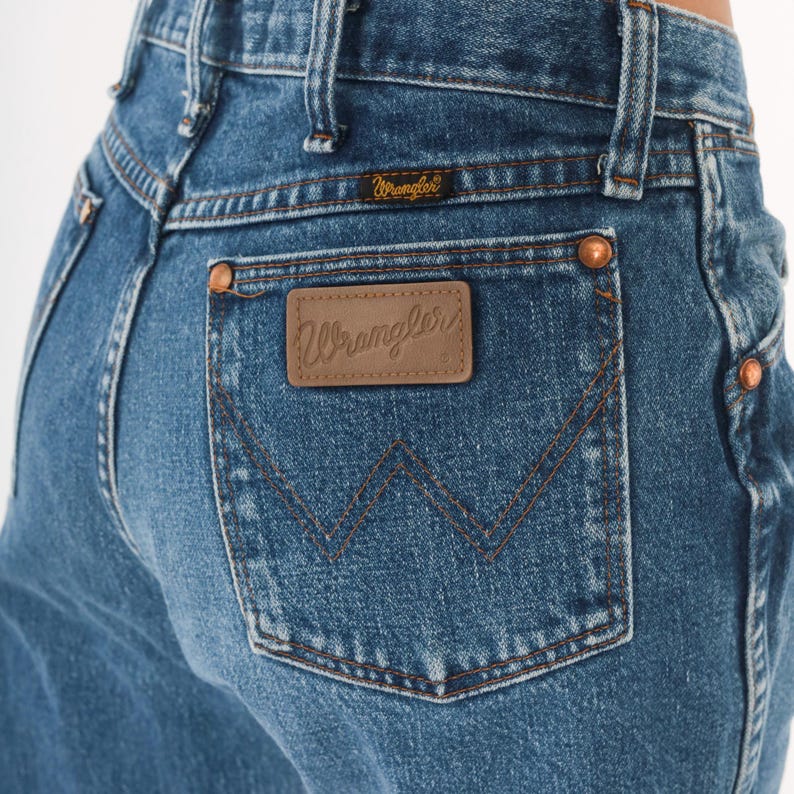 Puede incluir: Primer plano de unos vaqueros azules con una etiqueta Wrangler en el bolsillo trasero. Los vaqueros tienen un dise&ntilde;o cl&aacute;sico de cinco bolsillos con remaches de cobre y un parche de cuero marr&oacute;n. El logotipo de Wrangler es visible en el parche y encima del bolsillo trasero.