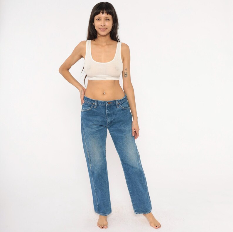 Pode incluir: Um top curto de cor clara e jeans azuis. Os jeans t&ecirc;m fecho de bot&atilde;o e um design cl&aacute;ssico de cinco bolsos. O denim &eacute; uma lavagem m&eacute;dia com algum desbotamento e desgaste.