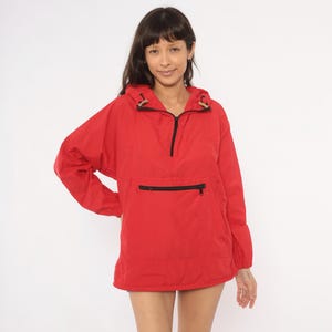May include: A bright red pullover windbreaker jacket featuring a hood with drawstring toggles, a half-zip front closure, and a large horizontal zippered pocket across the center. The jacket has long sleeves with elasticized cuffs and a straight hem.