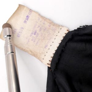 May include: A silver-colored stylus with a brown tip is positioned near a torn, aged paper tag. The tag is attached to a black fabric, possibly clothing. The paper has faded purple text.