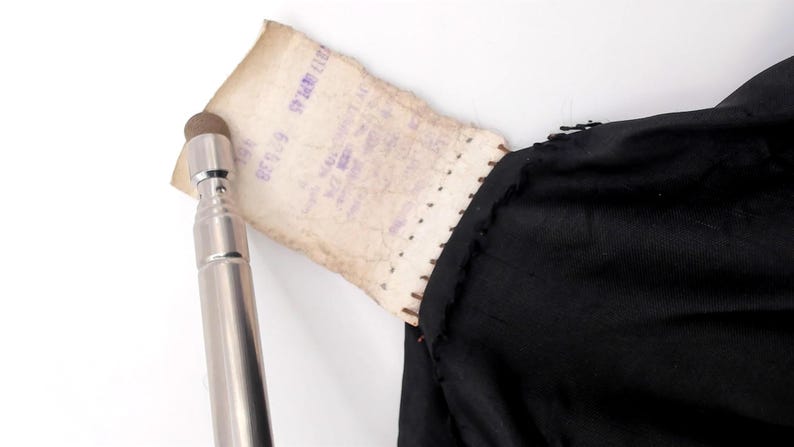 May include: A silver-colored stylus with a brown tip is positioned near a torn, aged paper tag. The tag is attached to a black fabric, possibly clothing. The paper has faded purple text.