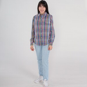 Pode incluir: Uma mulher vestindo uma camisa xadrez de manga comprida com cores azul, vermelho, verde e amarelo. Ela tamb&eacute;m est&aacute; vestindo jeans azul claro e t&ecirc;nis brancos.