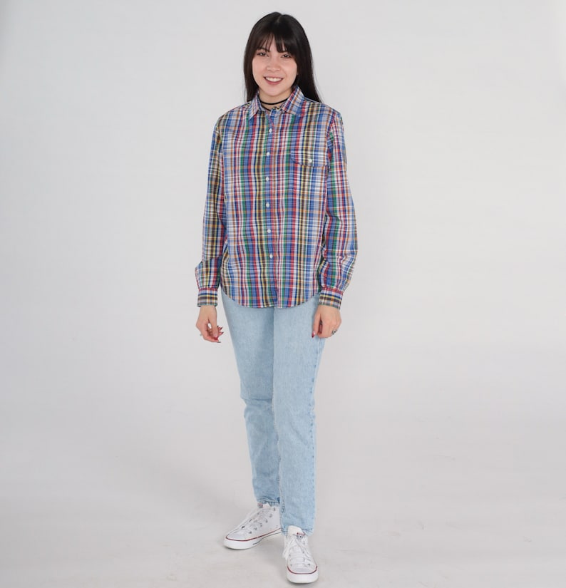 Pode incluir: Uma mulher vestindo uma camisa xadrez de manga comprida com cores azul, vermelho, verde e amarelo. Ela tamb&eacute;m est&aacute; vestindo jeans azul claro e t&ecirc;nis brancos.