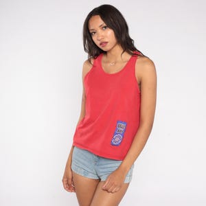 May include: A red tank top with a small patch that says "Bugle Boy" in blue and white. The tank top is sleeveless and has a scoop neck. The model is wearing light blue denim shorts.