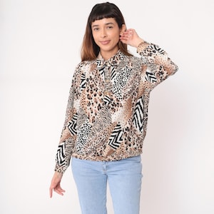 Pode incluir: Uma blusa de manga comprida com uma mistura de estampas de animais, incluindo manchas de leopardo e listras de zebra, em tons de marrom, bege e preto. A blusa tem gola e cintura franzida. A modelo está usando jeans azul claro.