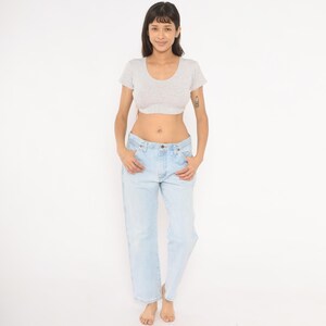 May include: A woman wearing a light gray crop top and light blue jeans. The jeans have a classic, relaxed fit. The top has a scoop neck and short sleeves. The woman is standing in front of a white background.