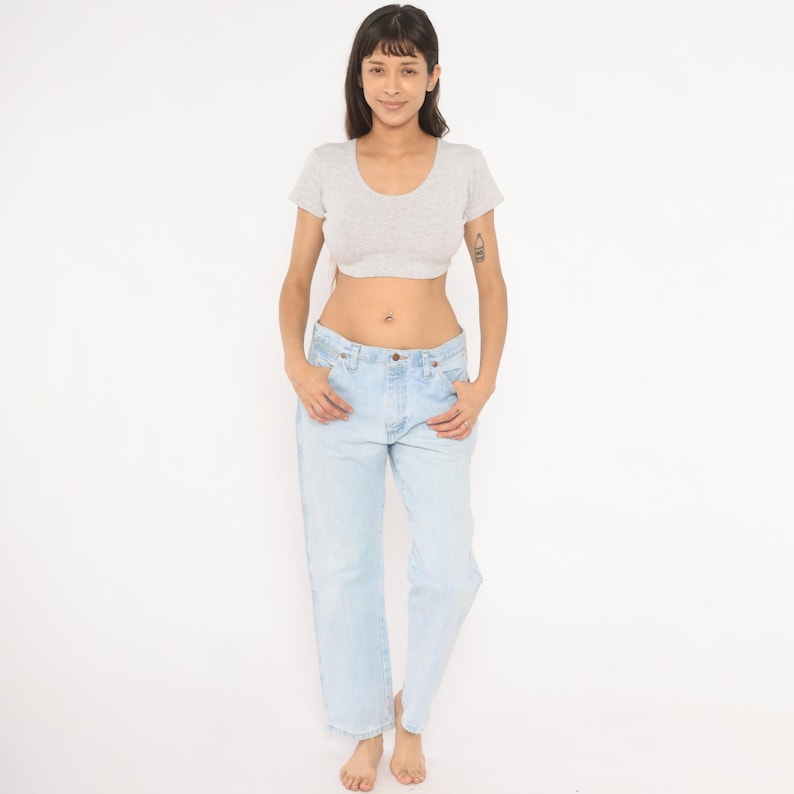 May include: A woman wearing a light gray crop top and light blue jeans. The jeans have a classic, relaxed fit. The top has a scoop neck and short sleeves. The woman is standing in front of a white background.