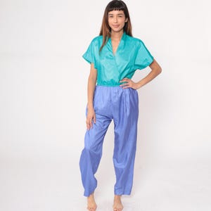 May include: A teal wrap-style top with short sleeves and high-waisted, light blue pants. The outfit has a shiny, silky appearance. The model is standing with one hand on their hip, showcasing the vintage style of the two-piece set.