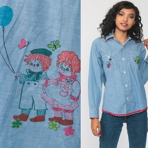 May include: A light blue button-down shirt with embroidered details. The shirt features two children holding balloons, a butterfly, and flowers. The shirt also has a red trim around the collar and cuffs.