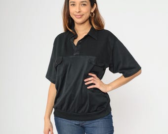 Puede incluir: Camisa negra de manga corta con cuello y dos bolsillos en el pecho. La camisa tiene una tapeta con botones y una cintura fruncida. La modelo lleva vaqueros azules.