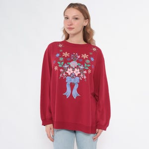 May include: A burgundy sweatshirt with a floral bouquet design and a blue bow. The floral arrangement includes pink, blue, and green elements. The long-sleeved top is made of a soft material.