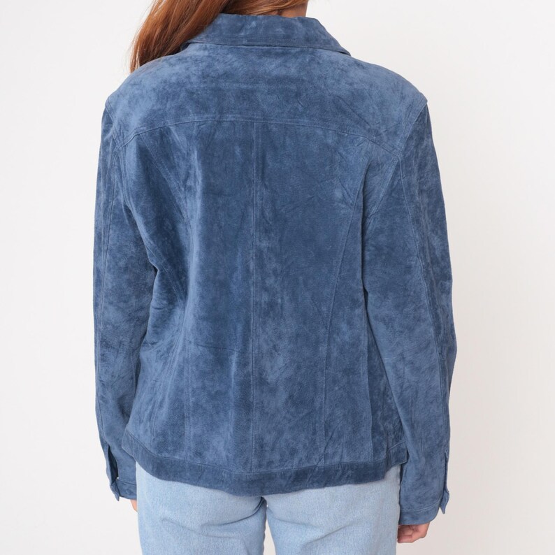 May include: A blue suede jacket with a button-down front and collar. The jacket has a classic design with a textured appearance. The sleeves are long, and the jacket is worn over light blue jeans. The jacket is a medium shade of blue.