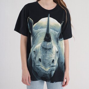 以下が含まれることがあります： 黒地に、サイの顔の大きなグラフィックプリントが施されたTシャツ。サイは灰色で、大きな角があります。