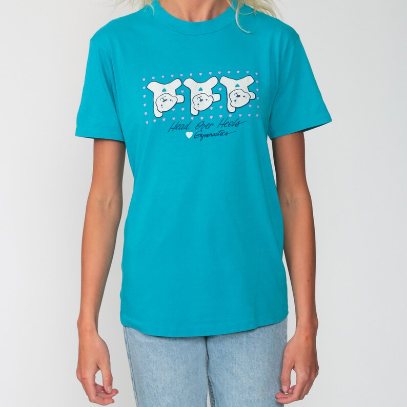 May include: A turquoise blue t-shirt with a graphic of three white bears doing a handstand. The text on the shirt reads "Head Over Heels Gymnastics".