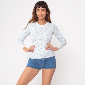 May include: A white long-sleeved top featuring a repeating light blue floral design. The top has a round neck and is made of a soft material. The top is worn with denim shorts.