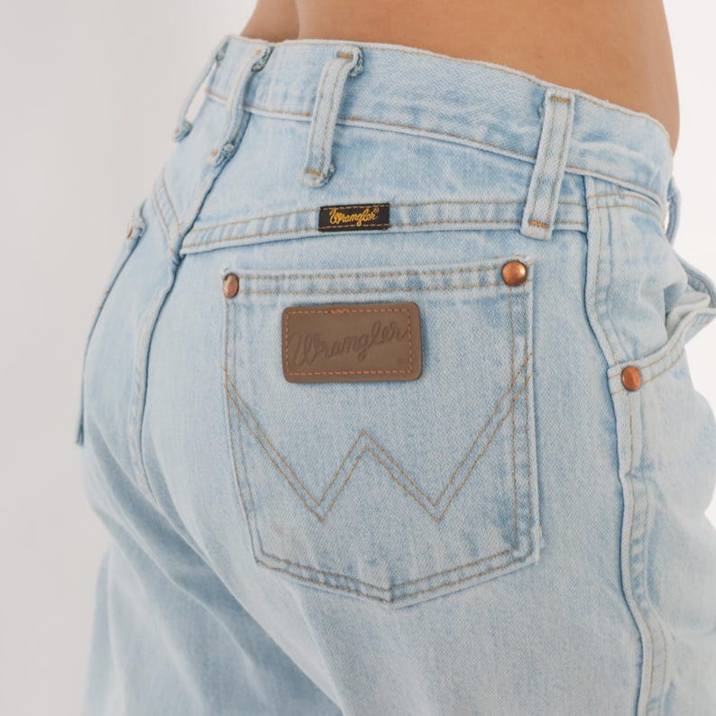 May include: Light blue denim jeans with the Wrangler logo on the back pocket and a small label above it. The jeans have copper-colored rivets and a classic five-pocket design. The jeans are a light wash.